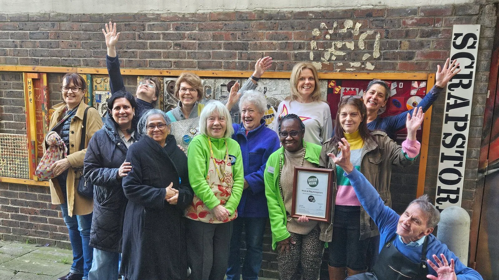 Scrapstore volunteers and staff celebrating outside the store on the Hazelhurst Estate, next to the hand-painted Scrapstore sign